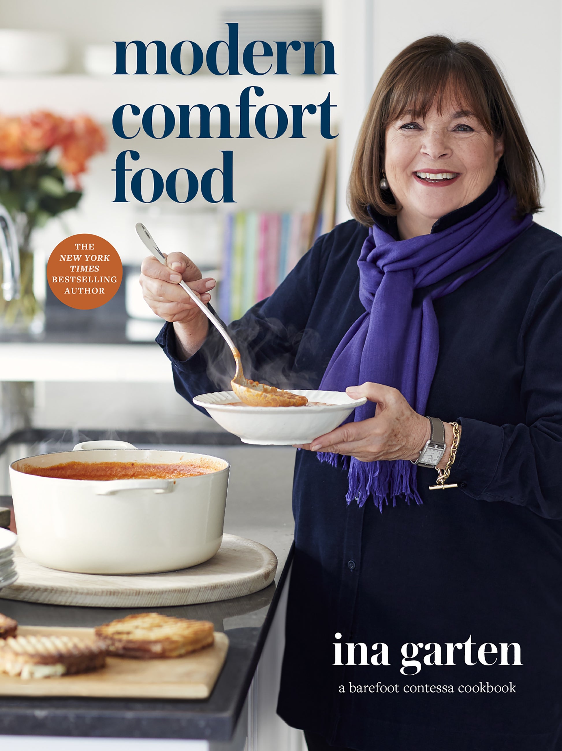 Ina Garten stands in a kitchen, holding a bowl of soup and grilled cheese, with a pot of soup on the stove. The cover features the product name “Modern Comfort Food by Ina Garten” and the brand name “Penguin Random House,” highlighting her signature collection of comforting food recipes.