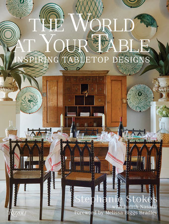 Book cover of "The World At Your Table" by Rizzoli, featuring a dining room with a wooden table, chairs, and decorative plates on the wall, showcasing stunning tablescapes enriched with exotic items collected from her travel photography adventures.