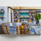 Open book featuring “Island Hopping” by Common Ground—pages reveal a living room with blue and white patterned decor, rattan furniture, bookshelves, plants, and a ship painting on the wall, capturing the relaxed elegance of Amanda Lindroth interiors inspired by island living.