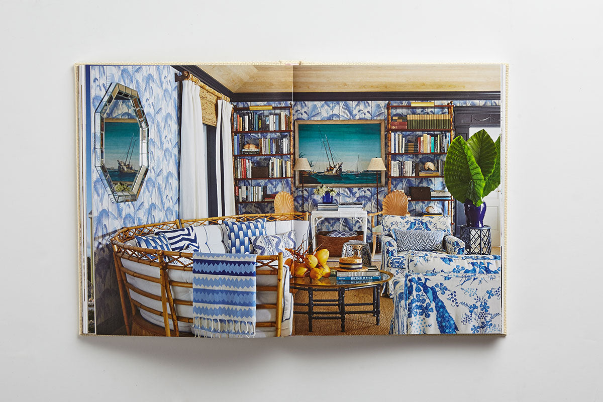 Open book featuring “Island Hopping” by Common Ground—pages reveal a living room with blue and white patterned decor, rattan furniture, bookshelves, plants, and a ship painting on the wall, capturing the relaxed elegance of Amanda Lindroth interiors inspired by island living.