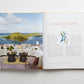 Open book displaying a seaside terrace overlooking the ocean and islands on the left page, evoking island lifestyle, with the title “Island Hopping” by Common Ground, accompanied by text and an illustration on the right page.