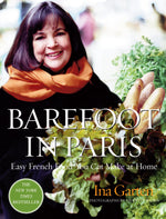 A woman with shoulder-length hair and an orange scarf smiles while holding a basket of fresh produce. She holds a copy of “Barefoot in Paris” by Penguin Random House, inviting readers to explore classic Parisian recipes.