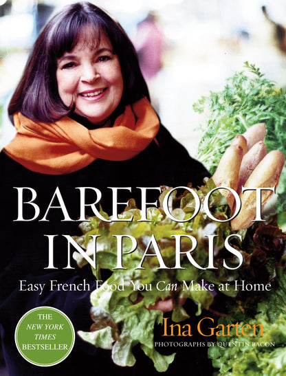 A woman with shoulder-length hair and an orange scarf smiles while holding a basket of fresh produce. She holds a copy of “Barefoot in Paris” by Penguin Random House, inviting readers to explore classic Parisian recipes.