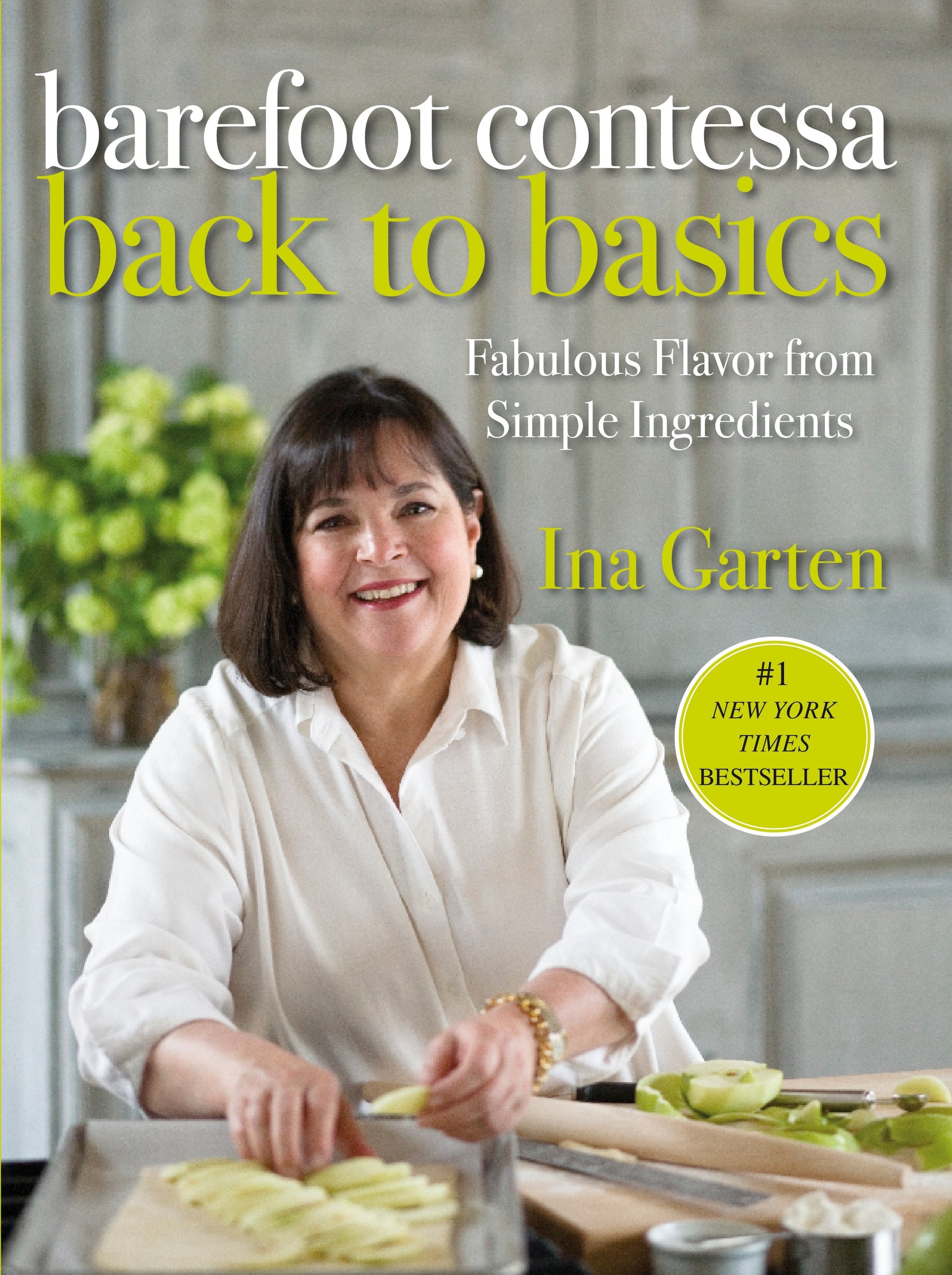 A woman with dark hair smiles while slicing apples in her kitchen, an open copy of “Barefoot Contessa Back To Basics” by Penguin Random House beside her—a perfect source of inspiration for easy home cooking.