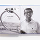 A black-and-white photo of tennis legend Arthur Ashe holding a racket spreads across two pages of the open book "US Open: 50 Years of Championship Tennis" by Abrams, celebrating iconic championship moments.