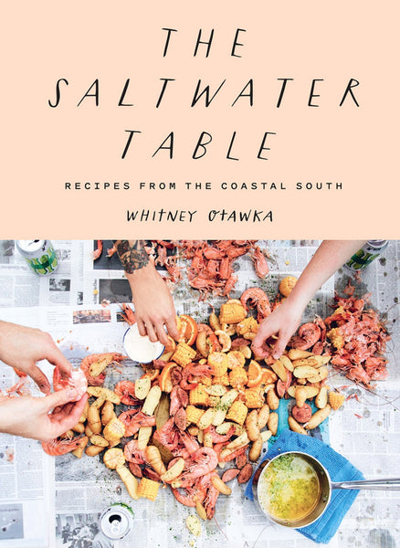 Hands gather around a coastal seafood boil with southern flavors, inspired by "The Saltwater Table: Recipes From The Coastal South" by Abrams. Spread on newspaper are shrimp, corn, and potatoes, alongside beverage cans and a steaming pot filled with local ingredients.