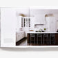 An open page of Atmosphere: The Seven Elements of Great Design showing a James Howard interior with a white kitchen, dark wood island, four metal stools, and pendant lighting.