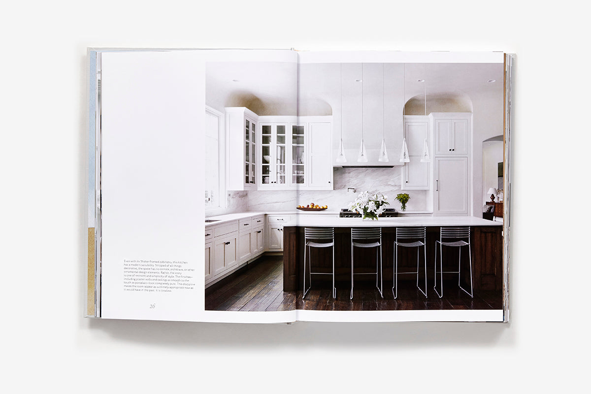 An open page of Atmosphere: The Seven Elements of Great Design showing a James Howard interior with a white kitchen, dark wood island, four metal stools, and pendant lighting.