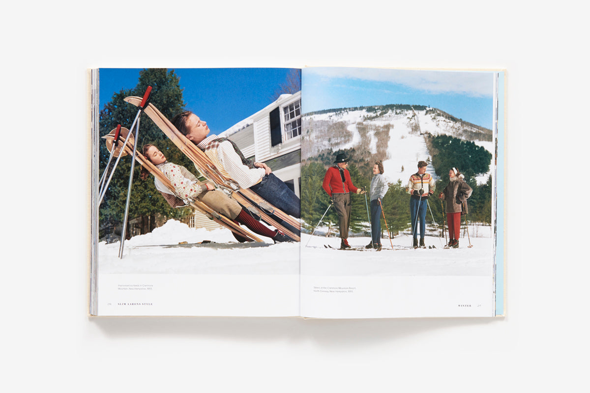 An open book from Abrams' "Slim Aarons: Style" shows glamorous personalities reclining on a sled on the left page and four people standing with skis against a snowy mountain backdrop on the right, capturing the essence of Slim Aarons' iconic fashion photography.