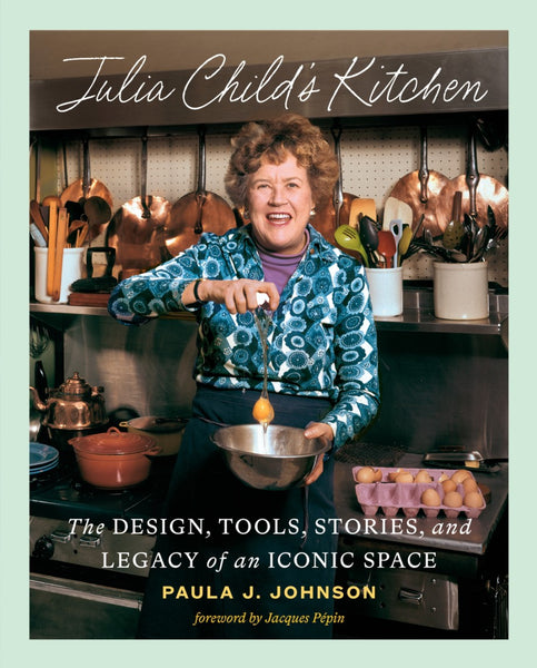 The book cover features a woman smiling while whisking in a copper-adorned kitchen, encapsulating the spirit of "Julia Child's Kitchen: The Design, Tools, and Stories" by Abrams. Home cooks will find it essential, with a foreword by Jacques Pépin linking past to present culinary practices.