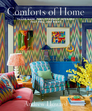 A vibrant living room with patterned wallpaper, bold furniture, abstract art, and a floral vase showcases Andrew Howard’s signature style. "Comforts of Home by Andrew Howard" from Abrams is featured in the scene.