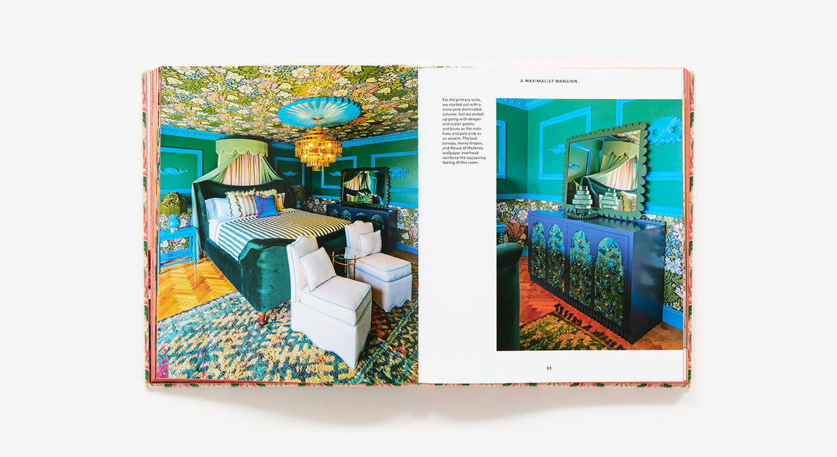 An open book of "The Maximalist: Colorful Interiors for Bold Living" by Abrams showcases a vibrant blue bedroom with patterned walls, ceiling, and carpet, ornate lighting, white chairs, and colorful home decor.
