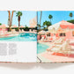 An open book, "The Maximalist: Colorful Interiors for Bold Living" by Abrams, displays a pastel pink poolside scene with lounge chairs, umbrellas, palm trees, and blue sky beside a page of text—offering vibrant home decor inspiration.