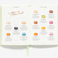 An open copy of "The Little Book of Ladurée: Patisserie" by Abrams shows descriptions and images of twelve colorful macarons, each pictured in a different shade against a white background.