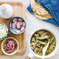 A saucepan filled with shredded meat in green sauce sits next to a tray of sliced radishes, pickled onions, shredded cabbage, flatbreads, and a blue checkered napkin—this colorful spread is inspired by recipes from *The Anti-Inflammation Cookbook* by Chronicle Books, crafted to make managing inflammation both delicious and enjoyable.