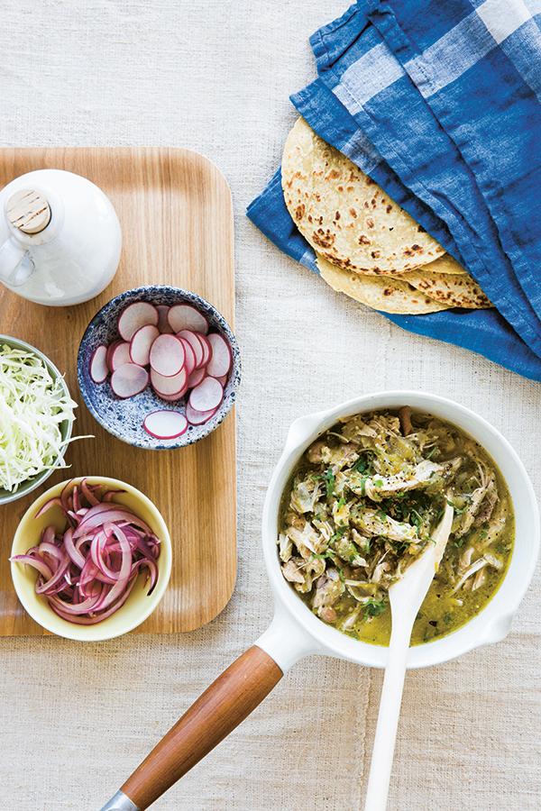 A saucepan filled with shredded meat in green sauce sits next to a tray of sliced radishes, pickled onions, shredded cabbage, flatbreads, and a blue checkered napkin—this colorful spread is inspired by recipes from *The Anti-Inflammation Cookbook* by Chronicle Books, crafted to make managing inflammation both delicious and enjoyable.