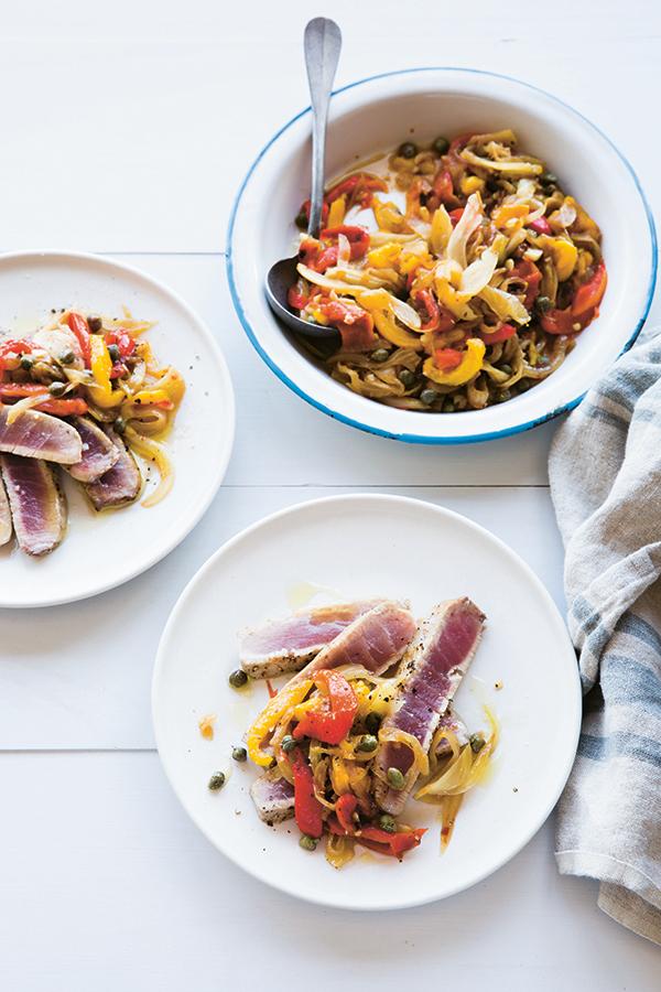 Two plates featuring seared tuna slices topped with sautéed peppers, onions, and capers create a vibrant meal inspired by recipes from The Anti-Inflammation Cookbook by Chronicle Books. Nearby, a generous serving bowl filled with the same inflammation-fighting dish and a spoon rests next to a striped cloth napkin for easy, family-style serving.