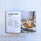 Open to *Platters and Boards* by Chronicle Books on a white table, displaying a recipe for “Breakfast in Bed” alongside a beautifully styled photo of a breakfast tray with juice, coffee, pastries, and fruit atop a black and white blanket—ideal inspiration for entertaining or finding fresh platter ideas.