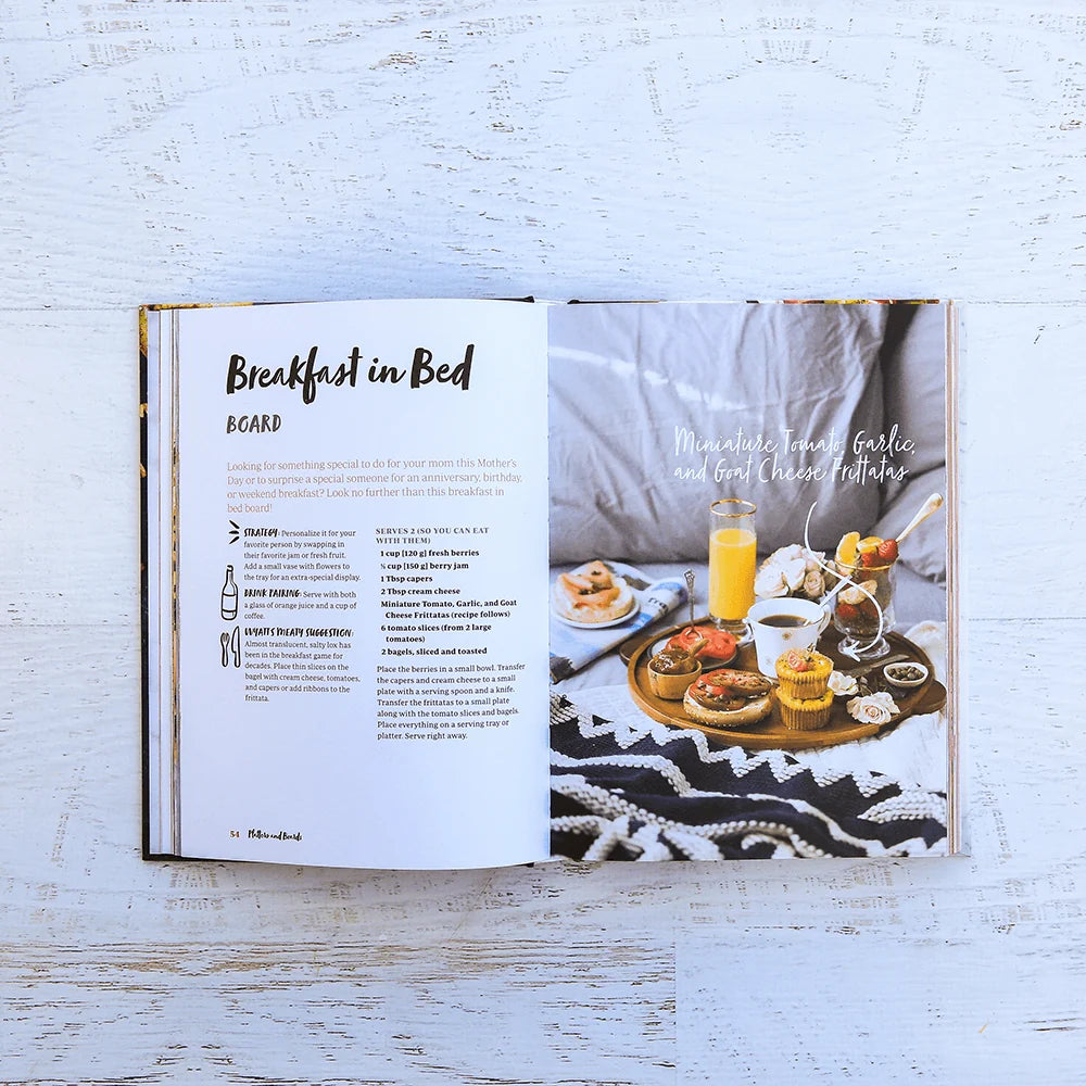 Open to *Platters and Boards* by Chronicle Books on a white table, displaying a recipe for “Breakfast in Bed” alongside a beautifully styled photo of a breakfast tray with juice, coffee, pastries, and fruit atop a black and white blanket—ideal inspiration for entertaining or finding fresh platter ideas.