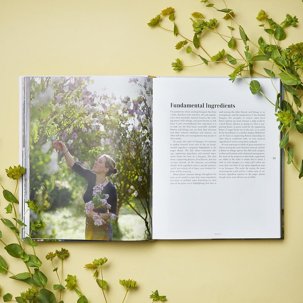 On a yellow surface adorned with green leafy sprigs lies A Year in Flowers by Chronicle Books, open to a spread that inspires creativity: the left page features an image of a woman gathering blossoms—an ideal scene for envisioning seasonal arrangements—while the right page, titled “Fundamental Ingredients,” provides essential guidance on bouquet design and flower care.