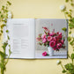 An open copy of “A Year in Flowers” by Chronicle Books displays a page of seasonal flower arrangement instructions on the left and a photo of a pink bouquet in a white vase on the right, framed by white flowers and greenery.