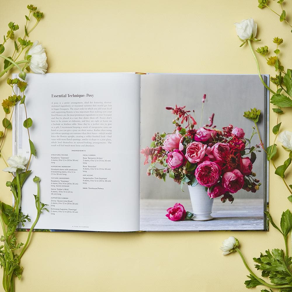 An open copy of “A Year in Flowers” by Chronicle Books displays a page of seasonal flower arrangement instructions on the left and a photo of a pink bouquet in a white vase on the right, framed by white flowers and greenery.