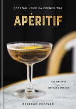 A coupe glass filled with a brown beverage, evoking the spirit of classic French apéritif culture, rests on a marble tabletop beside a book titled “Apéritif” from Penguin Random House.