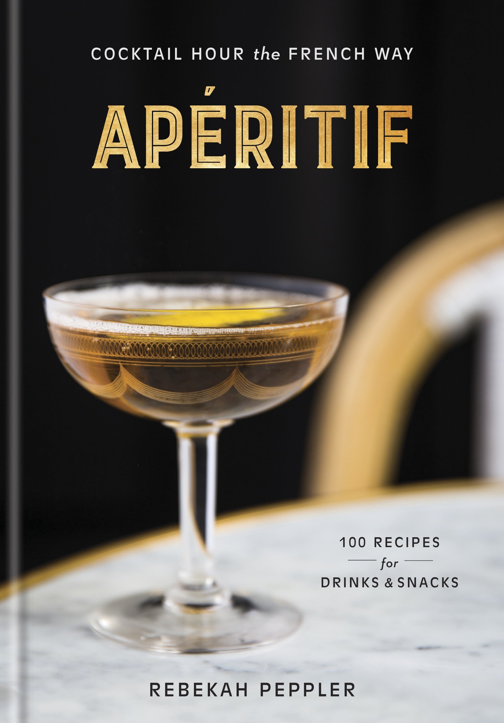 A coupe glass filled with a brown beverage, evoking the spirit of classic French apéritif culture, rests on a marble tabletop beside a book titled “Apéritif” from Penguin Random House.