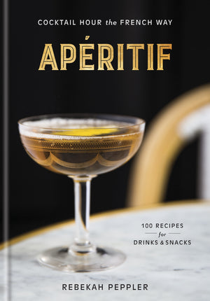 A coupe glass filled with a brown beverage, evoking the spirit of classic French apéritif culture, rests on a marble tabletop beside a book titled “Apéritif” from Penguin Random House.