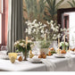 A dining table adorned with a white tablecloth is elegantly arranged with flowers, candles, pears, wine glasses, and plates in a room featuring expansive windows and a lush green plant mural—showcasing sophisticated home styling inspired by the approach found in Moments At Home from Chronicle Books.