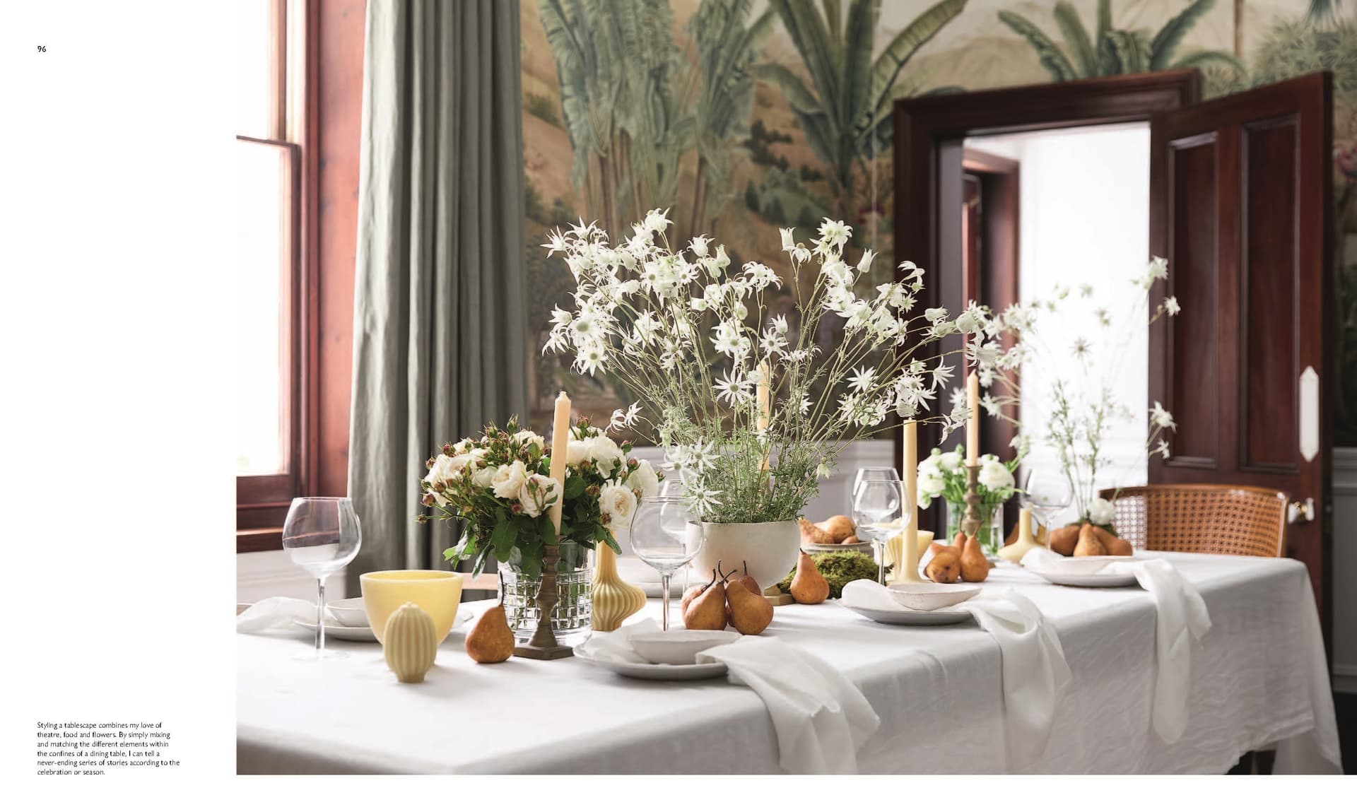 A dining table adorned with a white tablecloth is elegantly arranged with flowers, candles, pears, wine glasses, and plates in a room featuring expansive windows and a lush green plant mural—showcasing sophisticated home styling inspired by the approach found in Moments At Home from Chronicle Books.