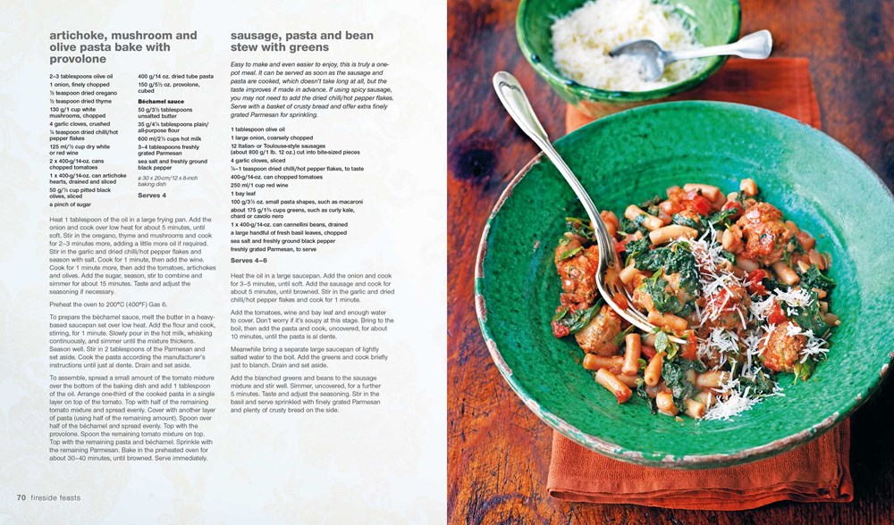 An open copy of *Comfort: A Winter Cookbook* by Common Ground rests under the warm glow of a kitchen light, displaying a cozy Fireside Feasts recipe. Nearby, a green bowl is filled with pasta and tomato sauce topped with cheese, ideal for those snug evenings indoors.