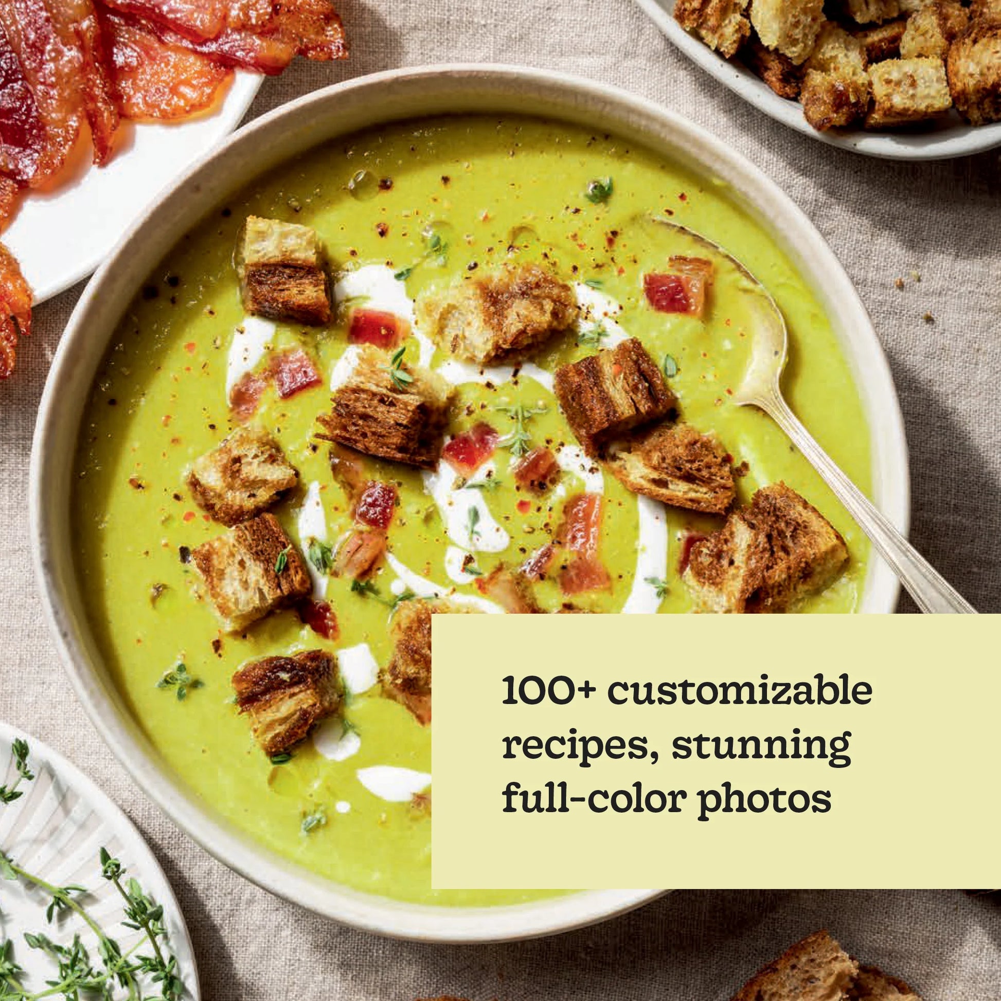 A bowl of vibrant green soup, garnished with golden croutons, creamy swirls, and crispy bacon, sits beside a plate of bacon and an extra serving of crunchy croutons—perfectly illustrating the effortless, customizable creations found in Chronicle Books’ *Every Season Is Soup Season*. With enticing photos and text emphasizing creative toppings for wholesome meals, this scene feels straight from your favorite healthy soup cookbook.
