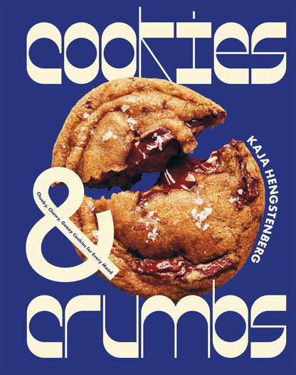 A large chocolate chip cookie broken in half sits on a blue background with the text “Cookies & Crumbs” and “Chronicle Books” displayed in bold, stylized fonts—perfectly capturing the irresistible appeal of gluten-free cookies.