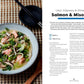 A bowl of udon noodles with salmon, edamame, shiitake mushrooms, and broth is placed next to black chopsticks and a spoon on a woven placemat, with the *Seriously Good Soups* cookbook by Chronicle Books open nearby—making for a truly comforting meal.