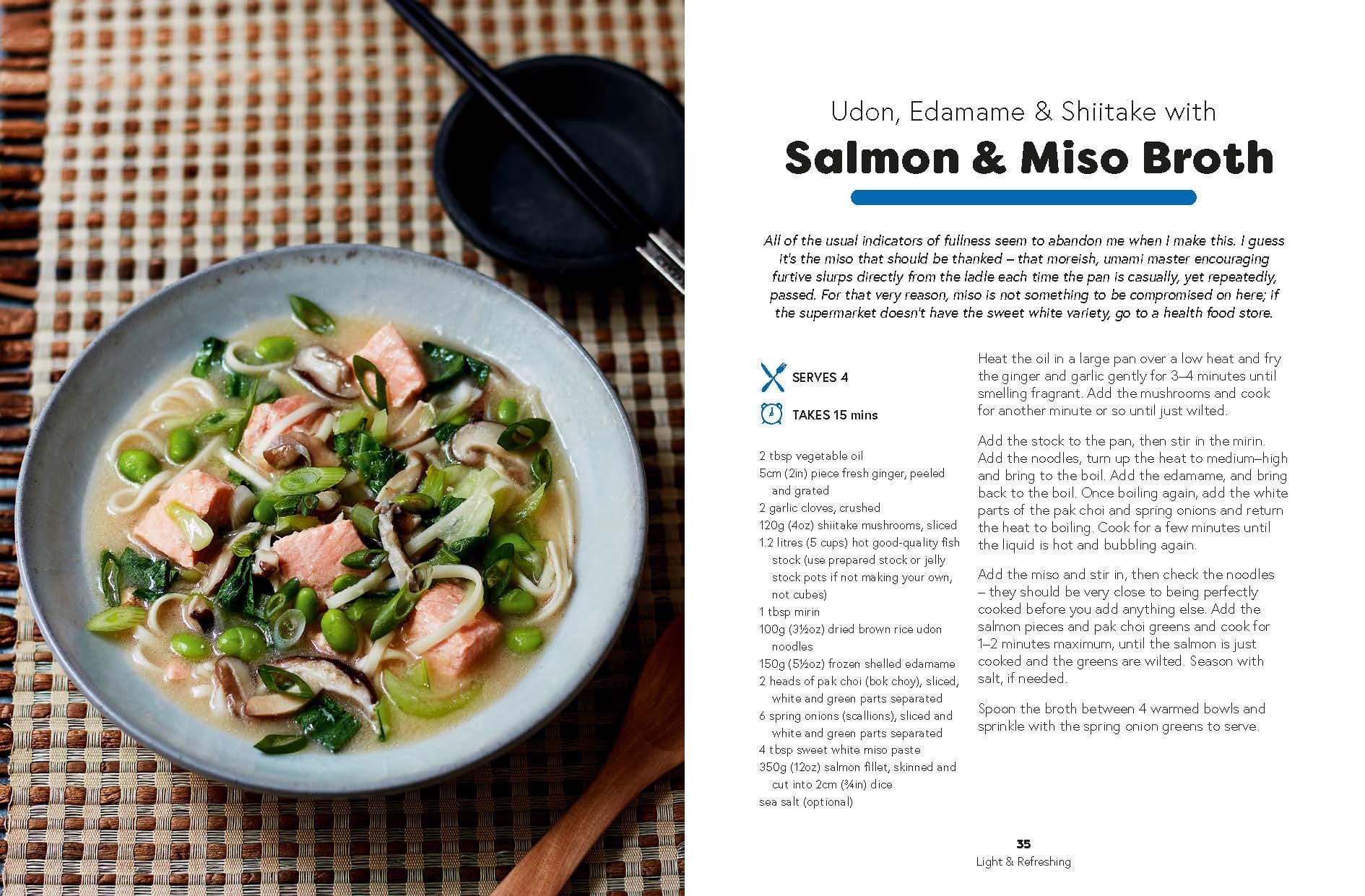 A bowl of udon noodles with salmon, edamame, shiitake mushrooms, and broth is placed next to black chopsticks and a spoon on a woven placemat, with the *Seriously Good Soups* cookbook by Chronicle Books open nearby—making for a truly comforting meal.