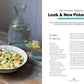 A bowl of leek and new potato soup with smoked haddock sits on a speckled plate, garnished with herbs; a spoon, bread, and a glass of water are nearby on a light table—an inviting scene inspired by nourishing recipes from *Seriously Good Soups* by Chronicle Books.