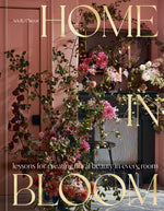 The cover of Home in Bloom by Penguin Random House features an elaborate floral arrangement displayed against a pink wall, with a candle and mirror in the background—an inspiring portrayal of flowers in interior design.