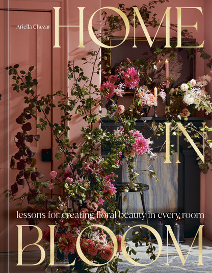 The cover of Home in Bloom by Penguin Random House features an elaborate floral arrangement displayed against a pink wall, with a candle and mirror in the background—an inspiring portrayal of flowers in interior design.