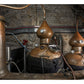 Two large copper pot stills with gauges and pipes sit in a stone-walled distillery room, surrounded by various equipment and red-striped cables—a classic scene reminiscent of traditional Scottish distilleries renowned for crafting exceptional Scotch from Penguin Random House.