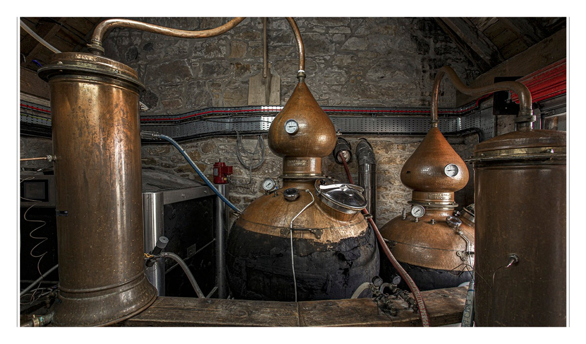 Two large copper pot stills with gauges and pipes sit in a stone-walled distillery room, surrounded by various equipment and red-striped cables—a classic scene reminiscent of traditional Scottish distilleries renowned for crafting exceptional Scotch from Penguin Random House.