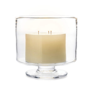The Nantucket Hurricane Collection by Simon Pearce features a three-wick white candle in a clear glass holder with a short pedestal base, inspired by classic trifle dishes.