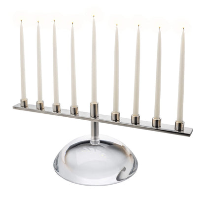 The Riverstone Menorah by Simon Pearce features a modern, handmade design with nine lit white taper candles on a reflective glass base, adding nature-inspired elegance to any space.