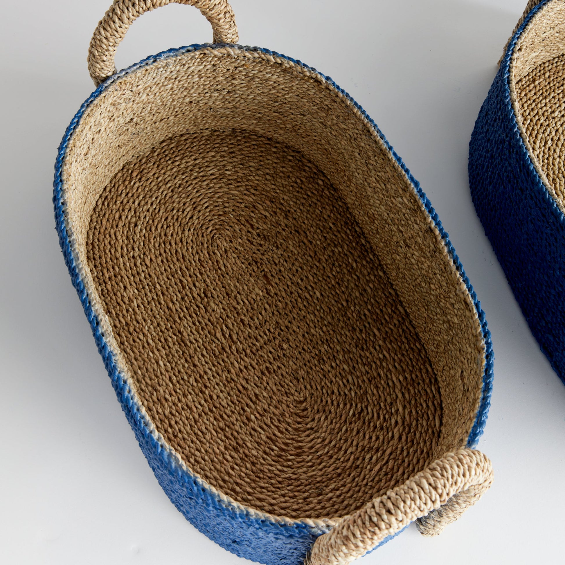 The Tiana Blue Seagrass Short Oval Basket Collection by Napa Home & Garden features a natural fiber interior, blue exterior, and rope handles—stylish and functional designer decor for any home.