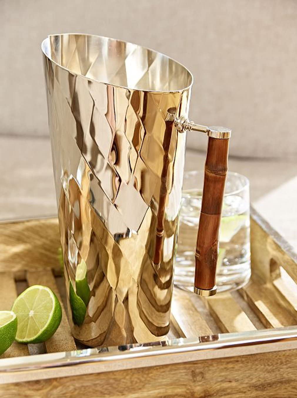 The ezrad Bamboo and Stainless Steel Decor Collection features a hammered stainless steel pitcher with a wooden handle, displayed on a tray beside lime wedges and an iced water glass, bringing modern elegance to any setting.