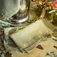 A stack of folded beige linen napkins from the Juliska Berry Scallop Napkin & Tablecloth Collection, featuring decorative trim, rests on a painted wooden surface alongside a metal pitcher, a glass vase with flowers, and coordinating woven linen tablecloths by Juliska.