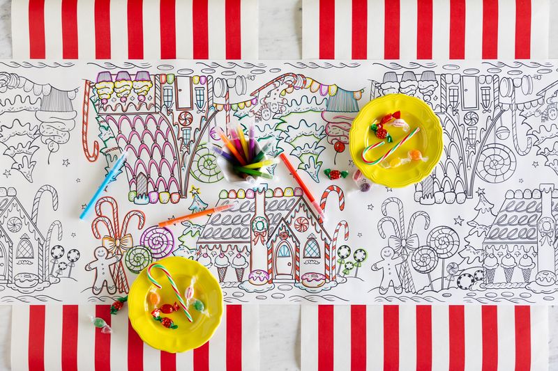 A Gingerbread Partyware Collection table runner with gingerbread house illustrations, markers, and two yellow plates of hard candies as festive accents.