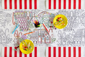 A Gingerbread Partyware Collection table runner with gingerbread house illustrations, markers, and two yellow plates of hard candies as festive accents.