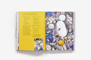 The Saltwater Table: Recipes From The Coastal South by Abrams opens to a yellow page of local ingredient recipes, paired with an overhead oyster roast table setting photo.