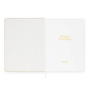 Sugar Paper’s Pale Pink Undated Weekly Planner features a striped inside cover and a title page embossed with “WEEKLY PLANNER” in gold lettering. A white ribbon bookmark keeps your place as you track habits and set goals each week.